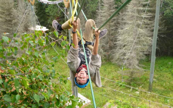 A person wearing safety gear and secured by ropes playfully hangs upside down on an obstacle on a high ropes course. 